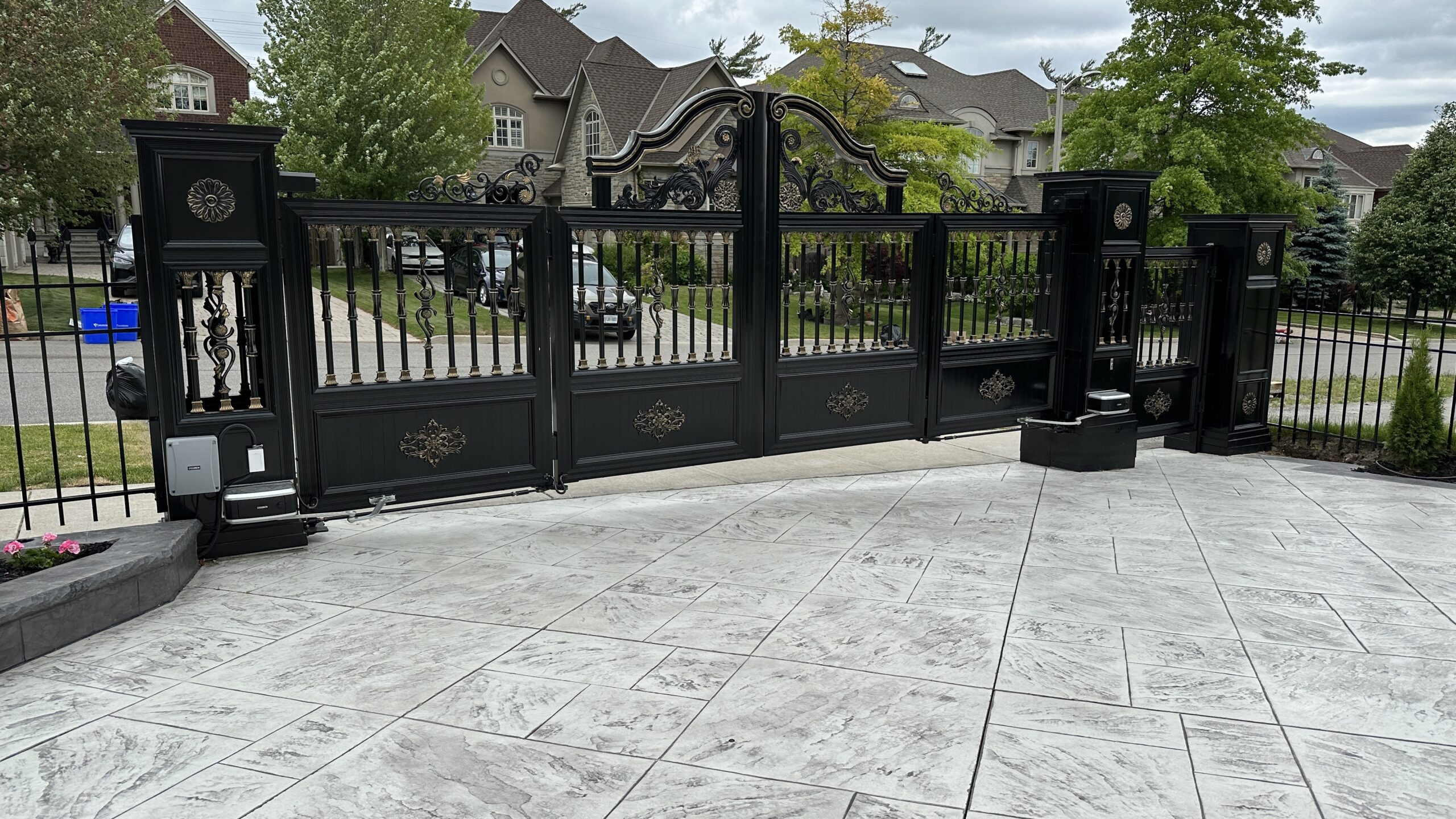 Closed black wrought-iron driveway gate in front of a suburban home. Closed black wrought-iron driveway gate in front of a suburban home.